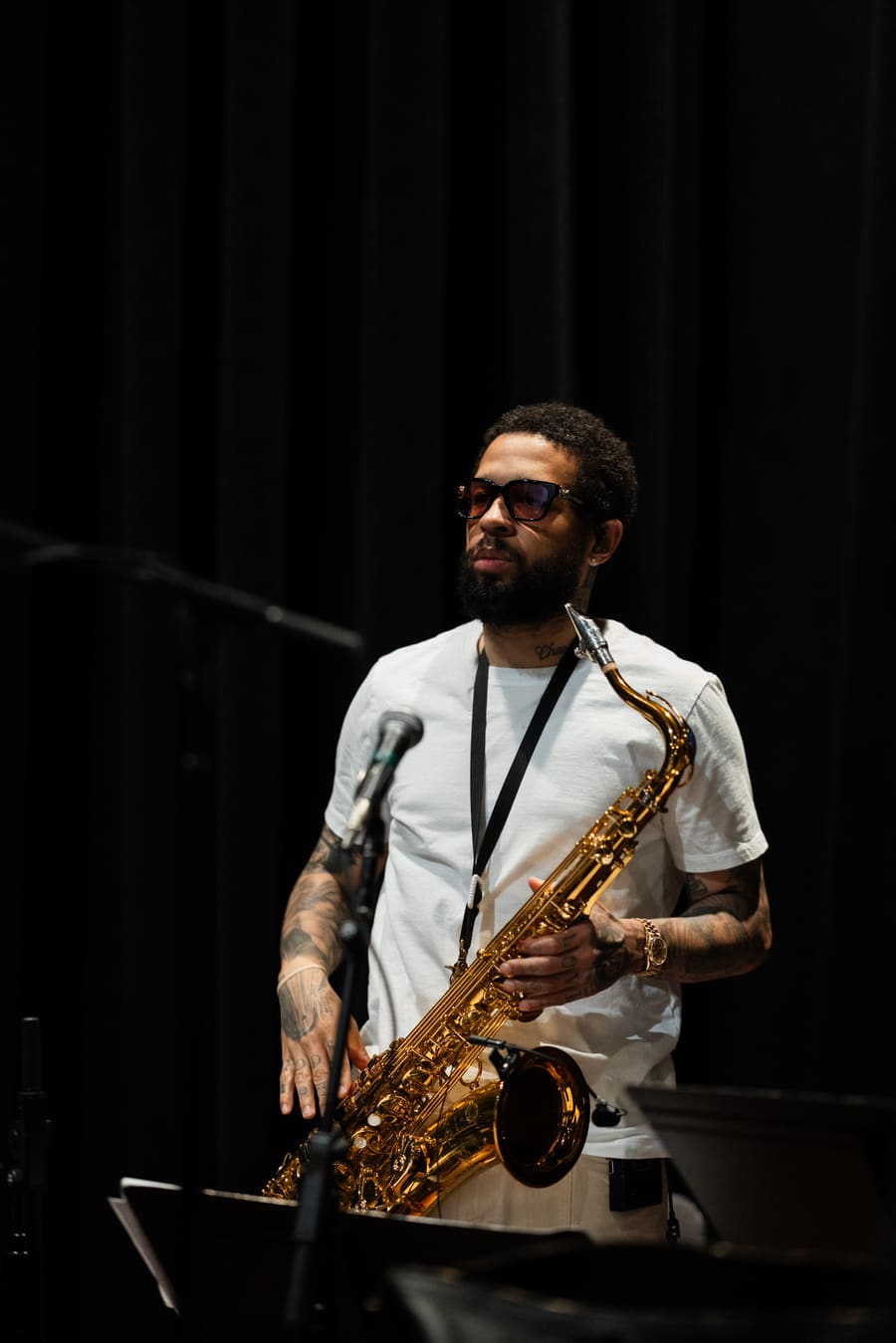 Kenneth Whalum in a white T-shirt with a tenor saxophone on a strap, in front of a black stage curtain